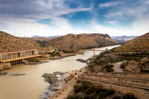 Gariep Damn wall, with water flowing through the dam gates into the river below, in a dry arid mountainous landscape covered in small shrubs, South Africa stock photo
