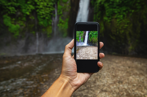 Male hand with a smartphone. Man taking picture of a beautiful waterfall.