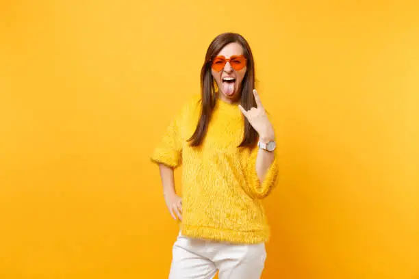 Funny crazy young woman in fur sweater and heart orange glasses screaming, showing rock-n-roll sign isolated on bright yellow background. People sincere emotions, lifestyle concept. Advertising area