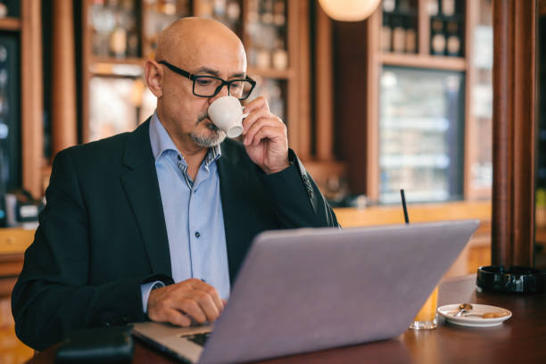 istock Bearded senior businessman in suit using laptop and drinking coffee while sitting in cafeteria. 1074619206