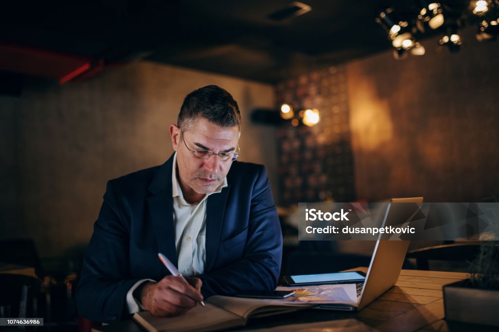 Middle-aged businessman with serious face writing in agenda while sitting in cafe at the evening. On table laptop, chart and tablet. Adult Stock Photo