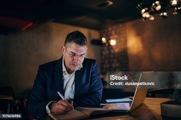 Middleaged Businessman With Serious Face Writing In Agenda While Sitting In Cafe At The Evening On Table Laptop Chart And Tablet Stock Photo - Download Image Now