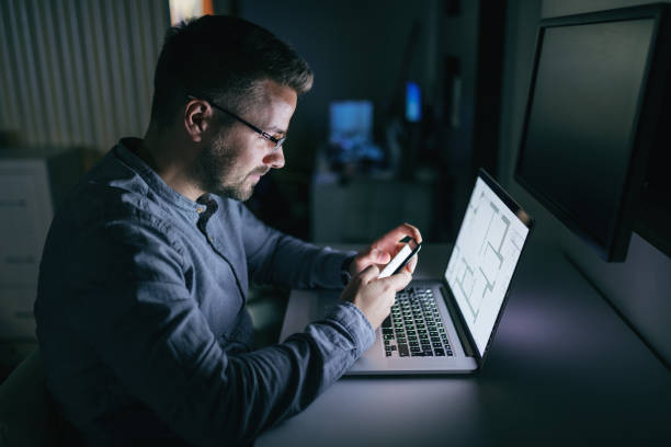 istock Young Caucasian businessman with eyeglasses using smart phone for writing or reading message while sitting in the office late at night. In front of him laptop. Side view. 1074638160