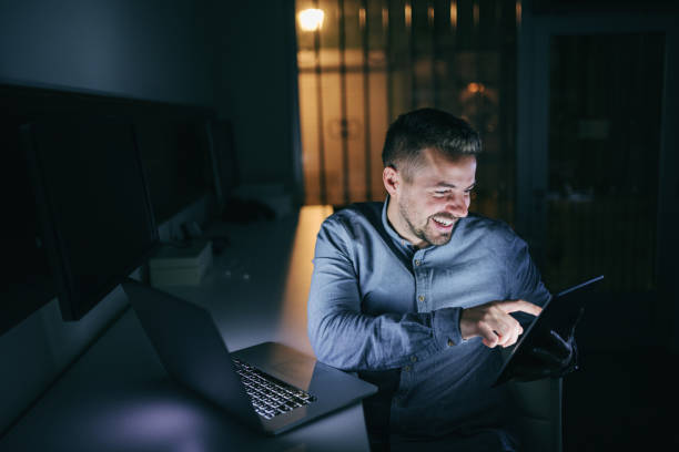 istock Young smiling Caucasian bearded employee using tablet while sitting in the office late at night. Next to him laptop. 1074639478