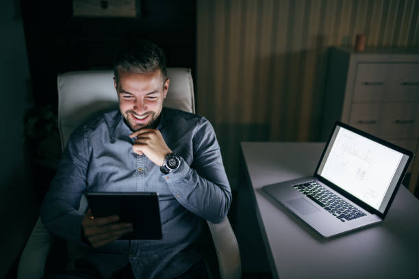 istock Young smiling Caucasian bearded employee using tablet while sitting in the chair late at night. Next to him desk with laptop. 1074639582