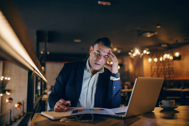 istock Concerned middle-aged businessman looking at chart and holding pen in hand while sitting in cafe at the evening. On the table coffee, laptop and agenda. 1074640412