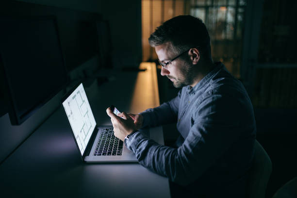 istock Young Caucasian businessman with eyeglasses using smart phone for writing or reading message while sitting in the office late at night. In front of him laptop. Side view. 1074642026