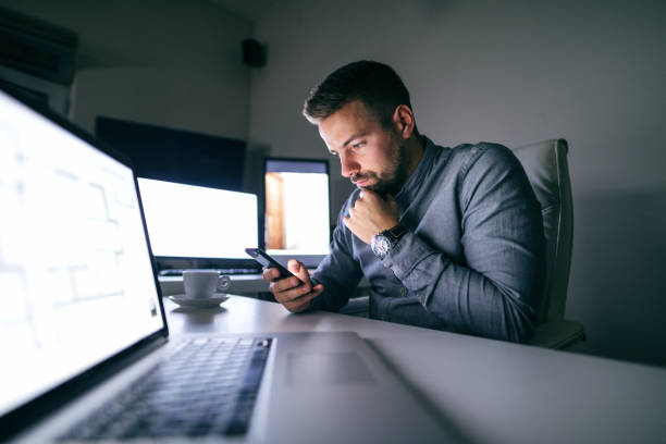 Thoughtful young Caucasian bearded employee reading or writing message on smart phone while sitting in the office late at night. On the desk laptop and cup of coffee. In background computer monitors. Selective focus on man.