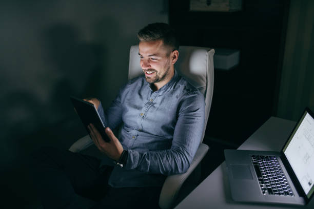 istock Young smiling Caucasian bearded employee using tablet while sitting in the chair late at night. Next to him desk with laptop. 1074647326