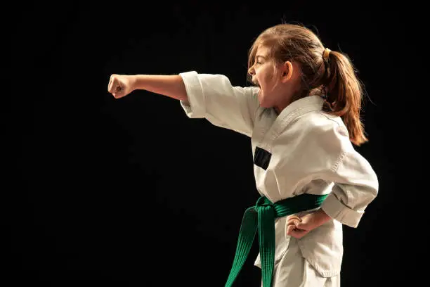 Little girl training martial arts against dark background. She wears white kimono with green belt