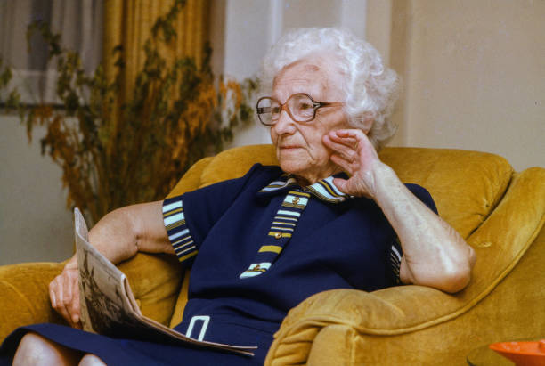 A silver haired female looks up from her magazine while sitting in her chair. She is looking off like she is in deep though and she is wearing wearing eye glasses and wears a blue dress.