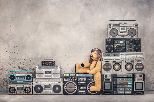 Teddy Bear toy with leather aviator's hat and goggles sitting on retro old school design ghetto blaster boombox stereo radio cassette tape recorders tower from circa 80s. Vintage style filtered photo