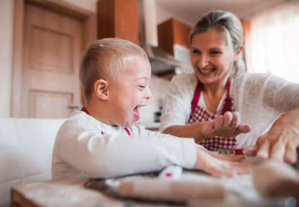 A laughing handicapped down syndrome child with his mother indoors baking. A laughing handicapped down syndrome child and his mother with checked aprons indoors baking in a kitchen, having fun. cleft lip stock pictures, royalty-free photos & images