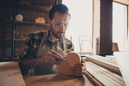 Close up photo portrait of serious concentrated focused bearded face looking down on small wooden heart in hand polishing the surface from thorns and splinters