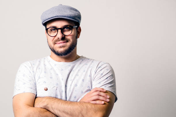 Confident man against gray background Portrait of confident creative businessman against white background. Male entrepreneur is smiling while standing with arms crossed. He is in smart casuals. flat cap stock pictures, royalty-free photos & images