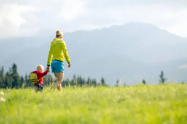 Happy mother with baby boy walking on green meadow. Hiking adventure with child on summer family trip in mountains. Vacations or weekend activity with child.