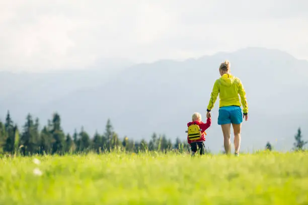 Happy mother with baby boy walking on green meadow. Hiking adventure with child on summer family trip in mountains. Vacations or weekend activity with child.
