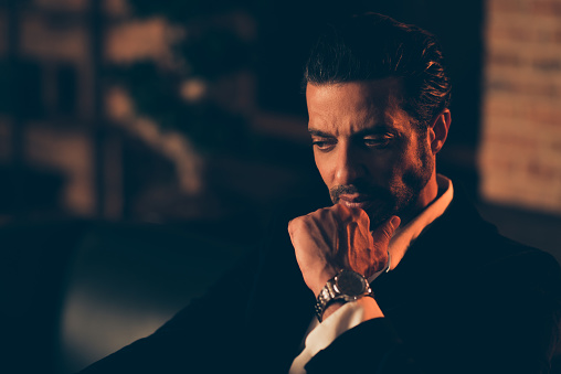 Half turn serious ambition focused calm stubble billionaire person man in elegant tuxedo and chic blazer indoor loft industrial interior look aside hold hand with watch on chin