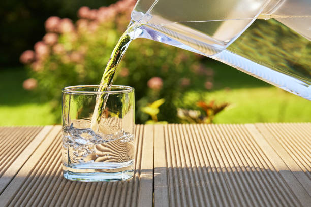 Pouring clear filtered water from a water filtration jug into a glass in green summer garden in a sunny summer day stock photo