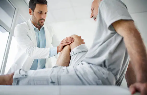 Closeup side view of an early 60's senior gentleman having some knee pain. He's at doctor's office having medical examination by a male doctor. The doctor is touching the sensitive area and trying to determine the cause of pain.