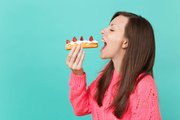 Side view of young woman in knitted pink sweater with closed eyes hold in hand, eating eclair cake isolated on blue turquoise background, studio portrait. People lifestyle concept. Mock up copy space vista lateral de una mujer joven en suéter hecho punto rosa con ojos cerrados sostenga en la mano, comiendo pastel éclair aislado sobre fondo azul turquesa, retrato de estudio. concepto de estilo de vida de las personas. se burlan de espacio de copia.