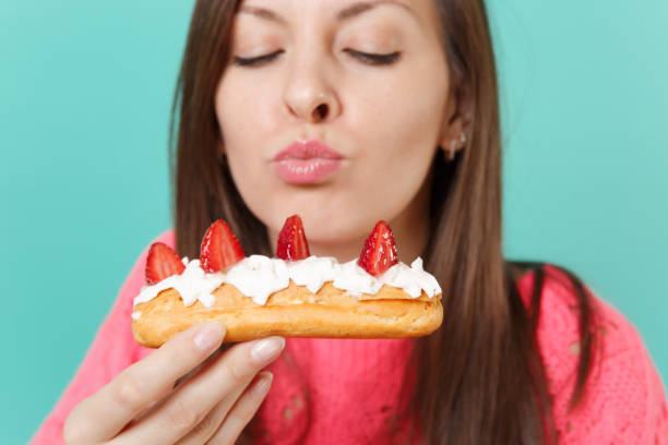 Cropped image of young woman in knitted pink sweater with closed eyes hold in hand, eating eclair cake isolated on blue wall background, studio portrait. People lifestyle concept. Mock up copy space imagen recortada de mujer joven en punto jersey rosa con cerró ojos sostenga en la mano, comiendo pastel éclair aislado sobre fondo de pared azul, retrato de estudio. concepto de estilo de vida de las personas. se burlan de espacio de copia.