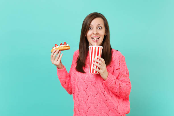 Excited young woman in knitted pink sweater holding in hands eclair cake, plastic cup of cola or soda isolated on blue wall background, studio portrait. People lifestyle concept. Mock up copy space joven emocionada en celebración de punto jersey rosa pastel éclair de manos, taza plástica de la cola o soda aislado sobre fondo de pared azul, retrato de estudio. concepto de estilo de vida de las personas. se burlan de espacio de copia.