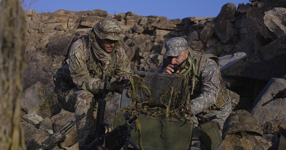 Medium shot of fully equipped and armed soldiers looking at a computer and another looking at a handheld device in the field and search coordinates