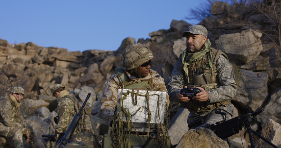 Medium shot of fully equipped and armed african american soldier looking at a computer and another talking on a radio and launches exploration drone