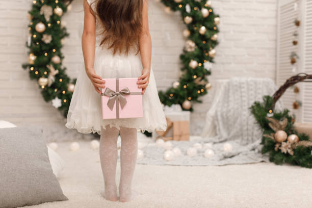 little child girl in white dress holding present box. Back view. child wants to put box with gift to the Christmas tree.