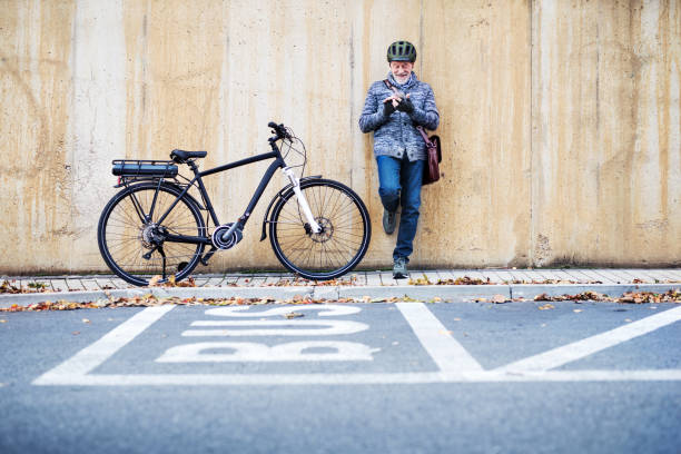 aktywny senior z rowerem elektrobikowym stojącym na zewnątrz w mieście, za pomocą smartfona. - cycling senior adult sports helmet men zdjęcia i obrazy z banku zdjęć