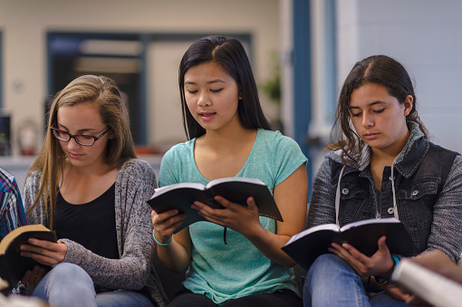 A group of high school students sit side by side in a classroom, reading from the bible.