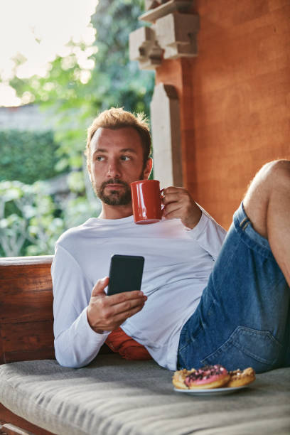 istock Man sitting on a terrace sofa and drinking coffee/tea, eating donuts while using cellphone. 1075007650