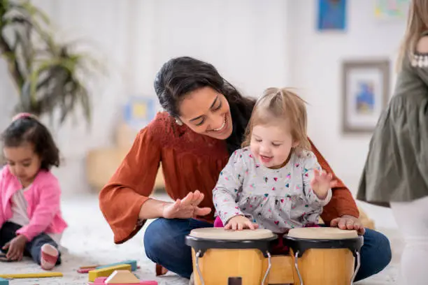 An adorable little girl sits in the lap of her daycare teacher. She is excited to learn how to play the bongos.
