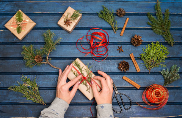 istock Gift wrapping. Packaging christmas present boxes. Top view of hands with fir tree, cedar, juniper, branches. Hand crafted gifts on blue wooden background. 1075035214