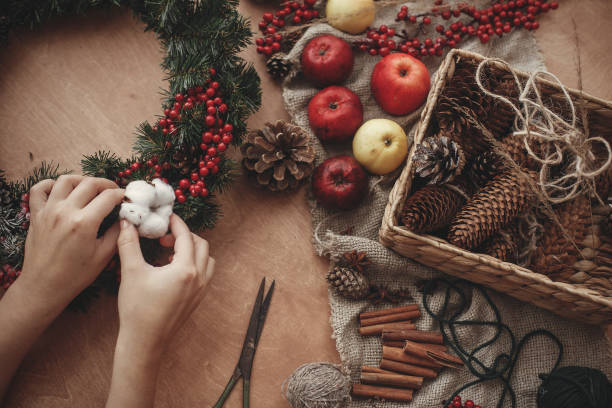 Rustic Christmas wreath. Hands putting cotton on fir branches, red berries and pine cones,thread, scissors, cinnamon on rustic wooden background. Atmospheric moody image at holiday workshop