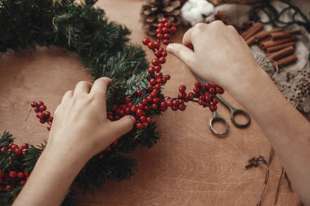 istock Rustic Christmas wreath. Hands holding fir branches, red berries and pine cones,rope, scissors, cinnamon, cotton on rustic wooden background. Atmospheric moody image at holiday workshop 1075036224