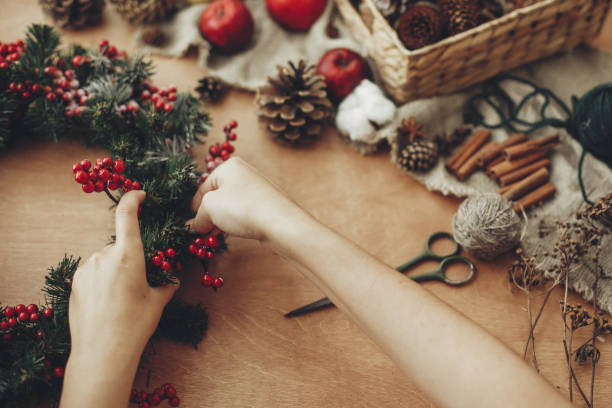 istock Hands making rustic christmas wreath with fir branches, red berries,pine cones,rope, scissors, cinnamon, cotton on rustic wooden background. Atmospheric moody image at holiday workshop 1075036302