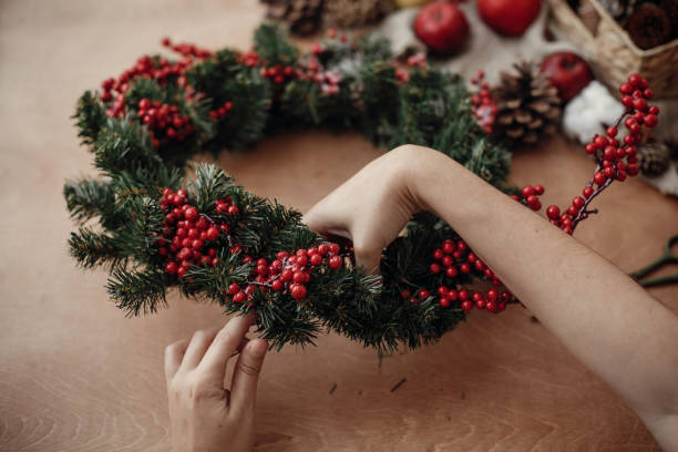 istock Rustic Christmas wreath. Hands holding fir branches, red berries and pine cones,rope, scissors, cinnamon, cotton on rustic wooden background. Atmospheric moody image at holiday workshop 1075036632