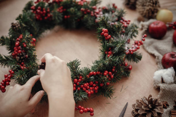 istock Rustic Christmas wreath. Hands holding fir branches, red berries and pine cones,rope, scissors, cinnamon, cotton on rustic wooden background. Atmospheric moody image at holiday workshop 1075036726