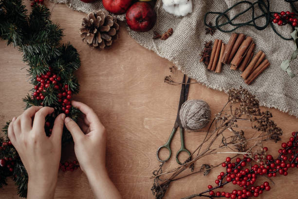 istock Rustic Christmas wreath flat lay. Hands holding fir branches, red berries and pine cones,thread, scissors, cinnamon, cotton on rustic wooden background. Making  wreath at workshop 1075037008