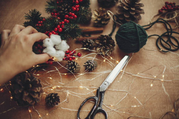 istock Hand holding cotton,pine cones, fir branches, wreath, red berries, thread, cinnamon,  lights on rustic wooden background. Details for making christmas wreath at workshop. Atmospheric image 1075037302