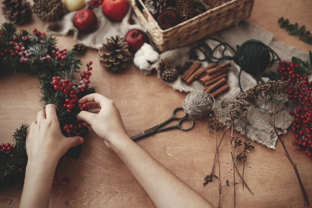 istock Rustic Christmas wreath. Hands holding fir branches, red berries and pine cones,rope, scissors, cinnamon, cotton on rustic wooden background. Atmospheric moody image at holiday workshop 1075037364