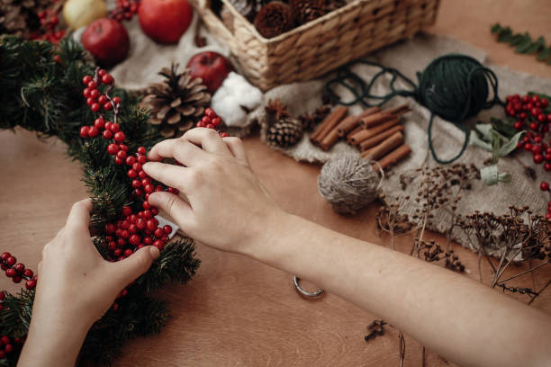 istock Hands making rustic christmas wreath with fir branches, red berries,pine cones,rope, scissors, cinnamon, cotton on rustic wooden background. Atmospheric moody image at holiday workshop 1075039182