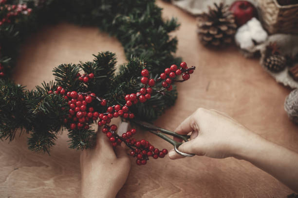 istock Hands making rustic christmas wreath, holding scissors at fir branches, red berries,pine cones,rope, cotton on rustic wooden background. Atmospheric moody image at holiday workshop 1075040058
