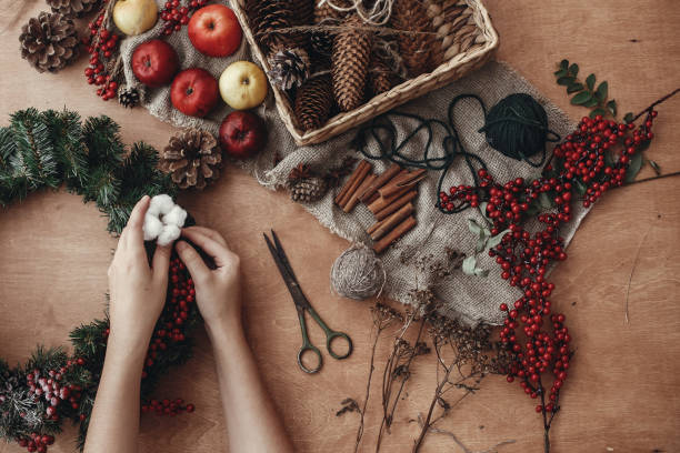 istock Rustic Christmas wreath flat lay. Hands putting cotton on fir branches, red berries and pine cones,thread, scissors, cinnamon on rustic wooden background. Making wreath at holiday workshop 1075040096