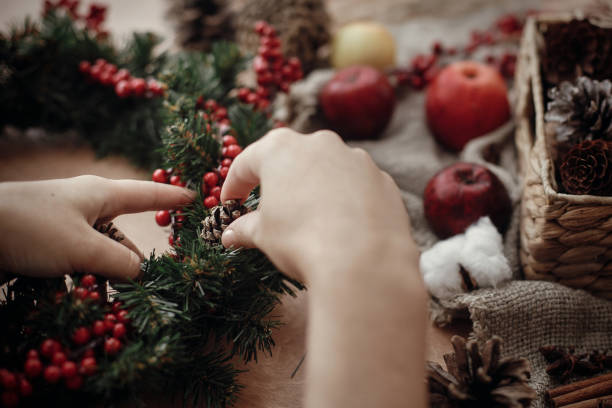 istock Rustic Christmas wreath. Hands holding fir branches, red berries and pine cones,rope, scissors, cinnamon, cotton on rustic wooden background. Atmospheric moody image at holiday workshop 1075040100