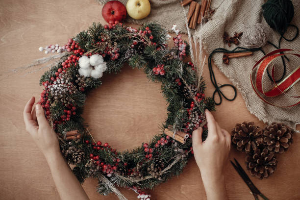 istock Rustic Christmas wreath flat lay. Hands holding fir branches, red berries and pine cones,thread, scissors, cinnamon, cotton on rustic wooden background. Making  wreath at workshop 1075044722