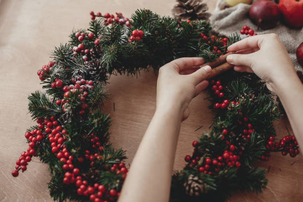 istock Hands making rustic christmas wreath, holding cinnamon at fir branches, red berries,pine cones,rope, scissors, cotton on rustic wooden background. Atmospheric moody image at holiday workshop 1075044928