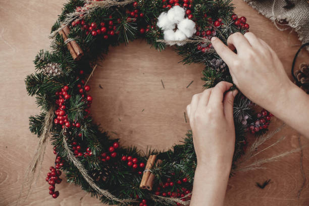 istock Hands making rustic christmas wreath, holding herbs at fir branches, red berries , pine cones, cotton on rustic wooden background. Atmospheric moody image at winter holiday workshop 1075044946